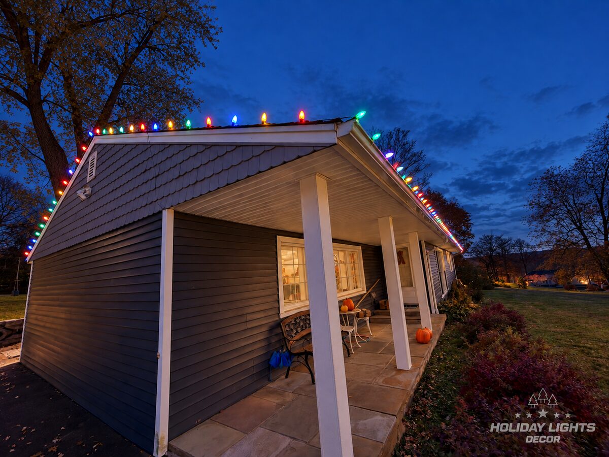 Covered porch and gable end outlined with multicolor C9 bulbs, seen from the driveway side of the ranch at dusk